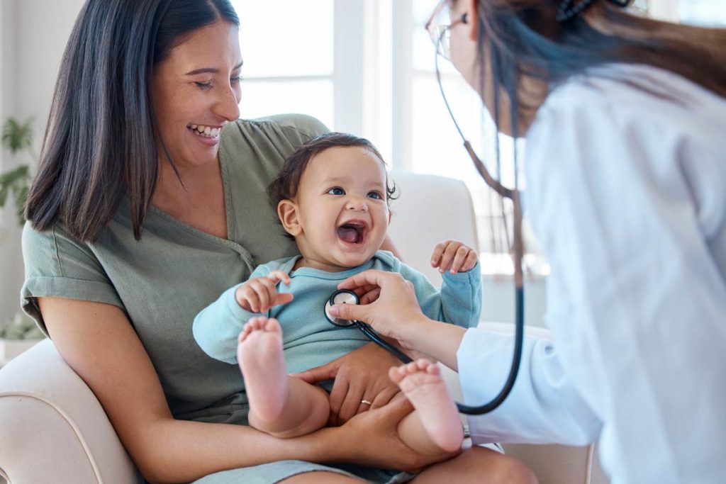 Baby laughing on mum's lap while female doctor uses stethoscope