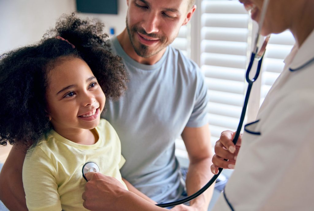 Young girl on man's lap while a doctor checks with a stethoscope 